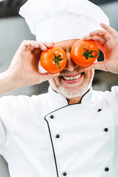 Chef masculino sonriente en uniforme y sombrero sosteniendo tomates en la cocina del restaurante - foto de stock