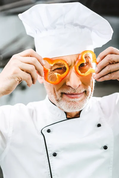 Chef masculino em uniforme e chapéu segurando fatias de pimenta na frente do rosto na cozinha do restaurante — Fotografia de Stock