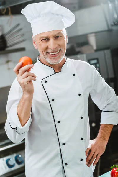 Souriant chef masculin en uniforme et chapeau tenant la tomate et regardant caméra dans la cuisine du restaurant — Photo de stock