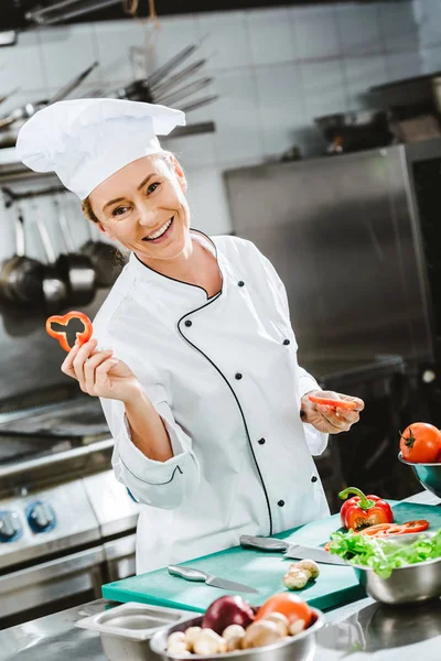 Lächelnde Köchin in Uniform mit Paprikascheiben und Blick in die Kamera beim Kochen in der Restaurantküche — Stockfoto