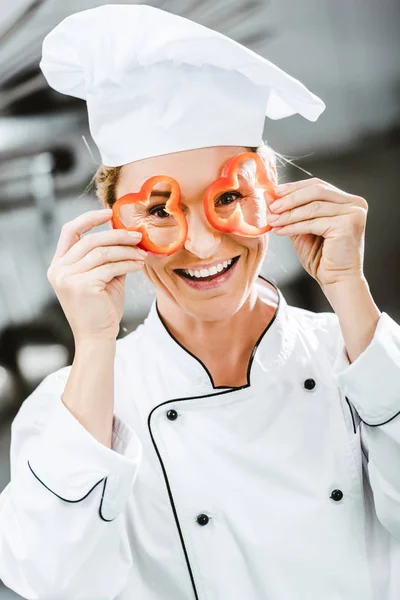 Chef sorridente in giacca doppiopetto con fette di pepe davanti al viso nella cucina del ristorante — Foto stock