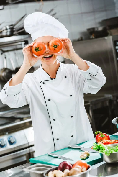 Femme chef souriante en veste à double boutonnage tenant des tomates devant le visage dans la cuisine du restaurant — Photo de stock