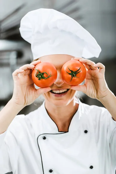 Smiling female chef in double-breasted jacket holding tomatoes in front of face in restaurant kitchen — Stock Photo