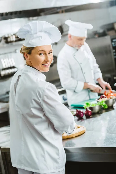 Belle chef féminine regardant la caméra et souriant tout en cuisinant avec un collègue dans la cuisine du restaurant — Photo de stock