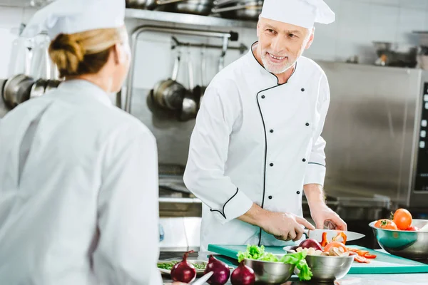 Foco seletivo de chefs masculinos e femininos em uniforme olhando uns para os outros enquanto cozinha comida na cozinha do restaurante — Fotografia de Stock