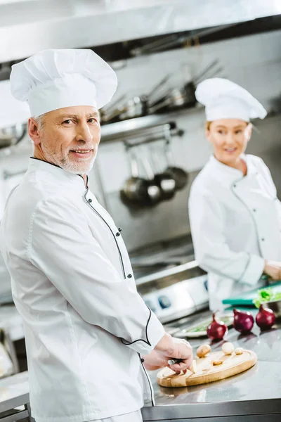 Guapo macho chef mirando cámara mientras cocinar con mujer colega en restaurante cocina - foto de stock