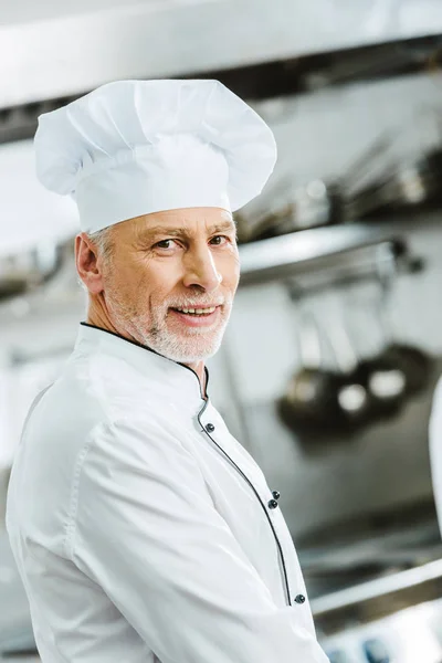 Guapo sonriente chef masculino en uniforme y gorra mirando a la cámara en la cocina del restaurante - foto de stock