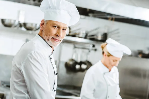 Selektiver Fokus eines gut aussehenden männlichen Kochs in Doppelbrustjacke und Hut, der in der Restaurantküche in die Kamera blickt — Stockfoto