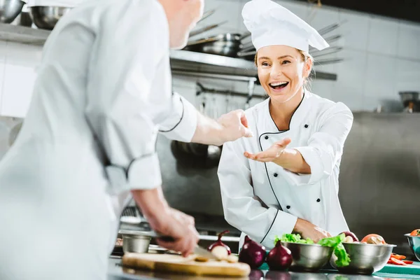 Smiling female chef in uniform taking ingredient from male colleague during cooking in restaurant kitchen — Stock Photo