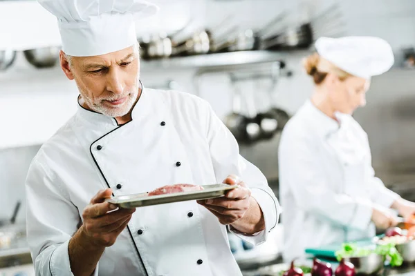 Chef masculino pensativo en uniforme sosteniendo plato de carne en el plato con colega en el fondo en la cocina del restaurante - foto de stock