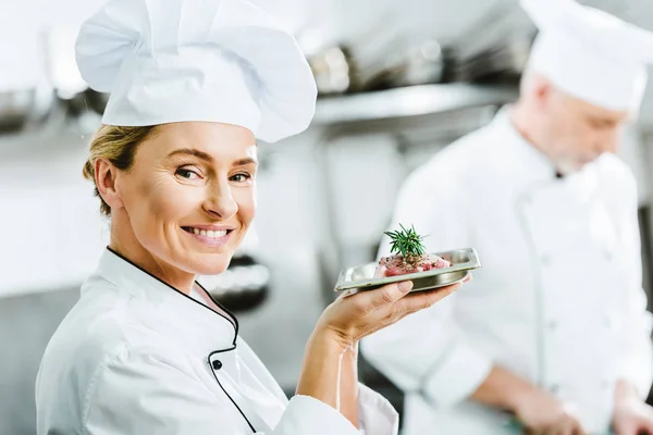 Beautiful smiling female chef in uniform holding plate with meat dish in restaurant kitchen and looking at camera — Stock Photo