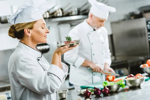 Chef femenino en uniforme sosteniendo plato de carne en el plato con colega de cocina masculina en el fondo en la cocina del restaurante - foto de stock