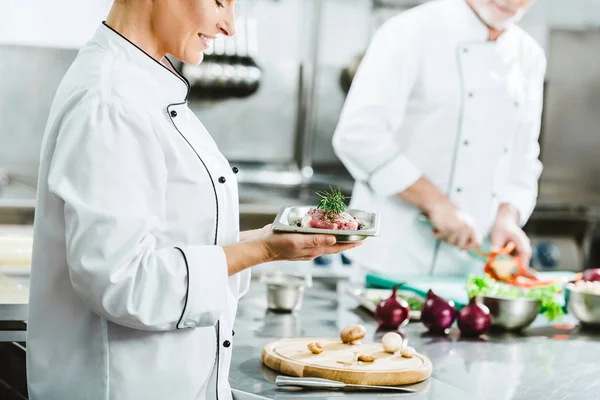 Cropped view of female chef in uniform holding meat dish on plate with colleague cooking on background in restaurant kitchen — Stock Photo