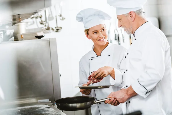 Chefs femeninos y masculinos en uniforme cocinando carne en sartén en la cocina del restaurante con espacio para copiar - foto de stock