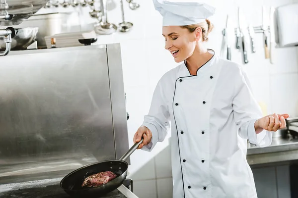 Beautiful smiling female chef in uniform cooking meat in restaurant kitchen — Stock Photo