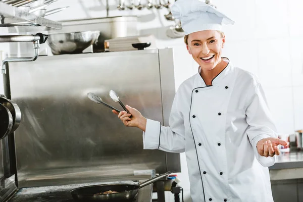 Beautiful female chef in uniform using kitchen tongs while cooking in restaurant kitchen — Stock Photo