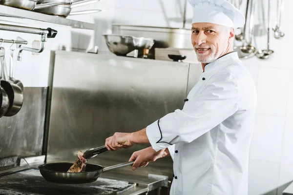 Chef masculino olhando para a câmera enquanto assando carne bife na cozinha do restaurante — Fotografia de Stock