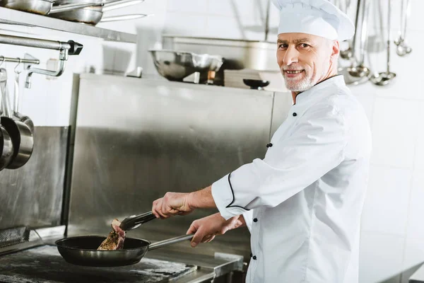 Mâle chef regardant caméra tout en rôtissant steak de viande dans la cuisine du restaurant — Photo de stock