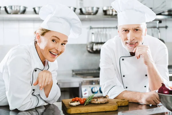 Chefs femeninos y masculinos felices en uniforme con filete de carne mirando a la cámara en la cocina del restaurante - foto de stock