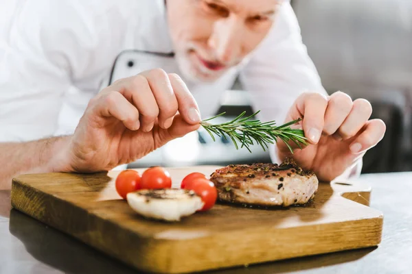 Nahaufnahme eines männlichen Küchenchefs in Uniform, der in der Restaurantküche Fleischsteak mit Rosmarin verziert — Stockfoto