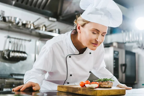 Beautiful female chef in uniform enjoying aroma of meat steak on wooden board in restaurant kitchen — Stock Photo