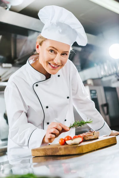 Hermosa cocinera en uniforme decorando filete de carne con romero en la cocina del restaurante - foto de stock