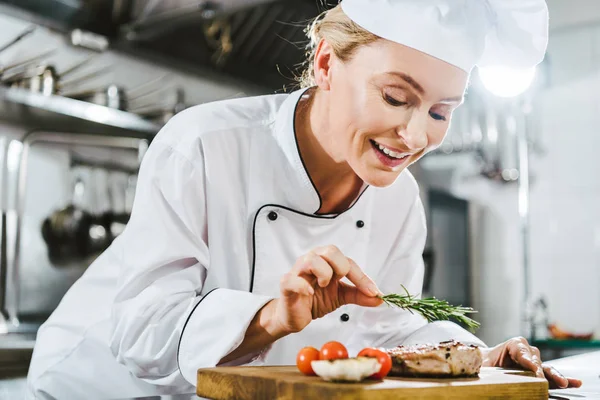 Hermosa cocinera en uniforme decorando filete de carne con romero en la cocina del restaurante - foto de stock