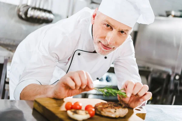 Männlicher Koch in Uniform dekoriert Fleischsteak mit Rosmarin und blickt in Restaurantküche in die Kamera — Stockfoto