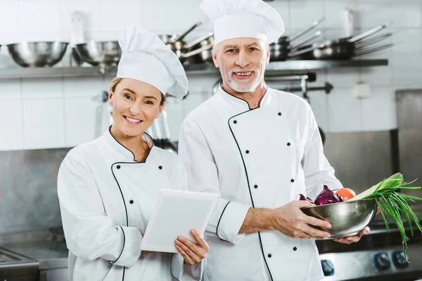 Chefs femeninos y masculinos en iniformes mirando a la cámara y usando tableta digital mientras cocinan en la cocina del restaurante - foto de stock