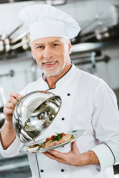 Schöner männlicher Koch in Uniform schaut in die Kamera und hält Kuppel vom Serviertablett mit Fleischplatte in der Restaurantküche — Stockfoto