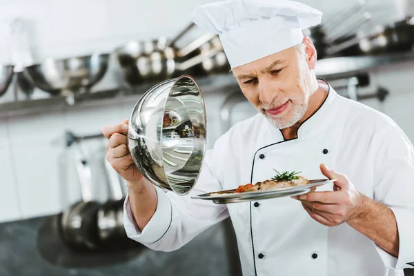 Enfoque selectivo de guapo chef masculino en uniforme de la celebración de la cúpula de bandeja de servir con plato de carne en la cocina del restaurante — Stock Photo