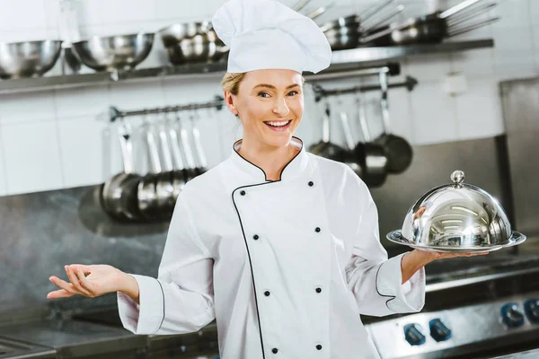 Красивая улыбающаяся женщина-шеф-повар в форме держит подающий поднос с куполом в кухне ресторана — стоковое фото
