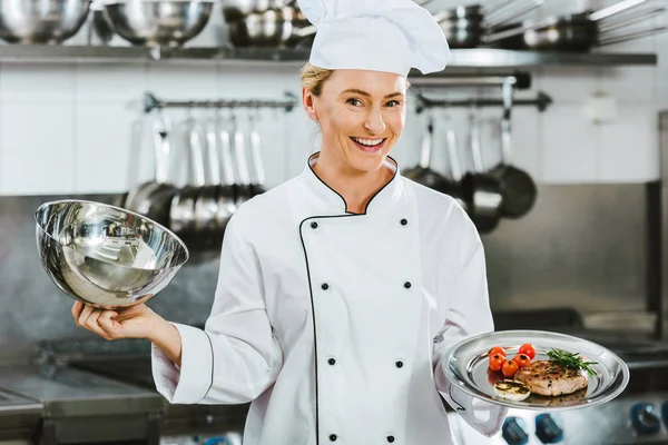 Schöne lächelnde Köchin in Uniform, die in der Restaurantküche Tablett mit Fleischplatte hält — Stockfoto