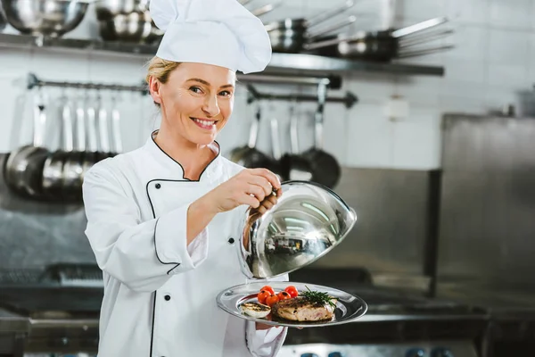 Beautiful smiling female chef in uniform holding dome from serving tray with meat dish in restaurant kitchen — Stock Photo