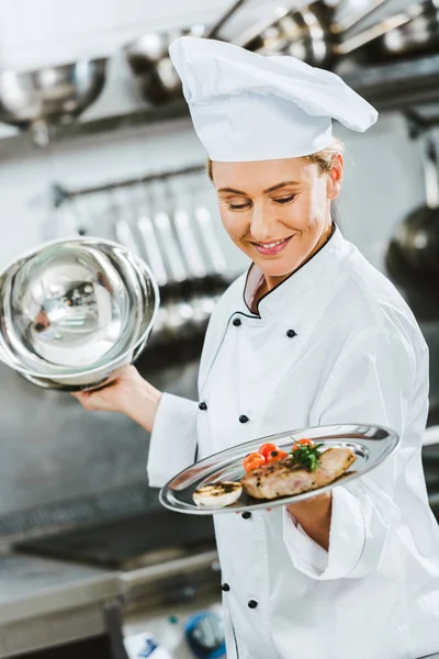 Selective focus of beautiful female chef in uniform holding serving tray with meat dish in restaurant kitchen — Stock Photo