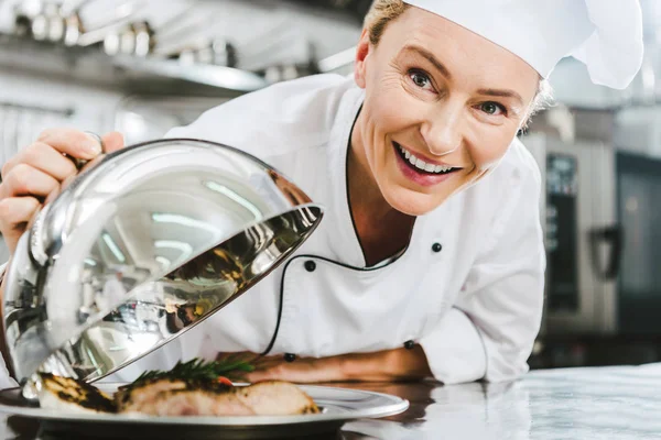 Beautiful female chef in uniform looking at camera and holding dome from serving tray with meat dish in restaurant kitchen — Stock Photo