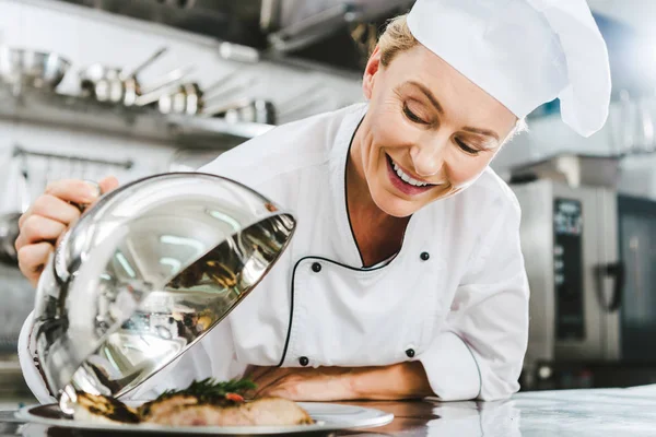 Beautiful female chef in uniform holding dome from serving tray with meat dish in restaurant kitchen — Stock Photo