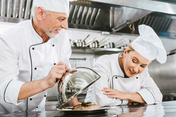 Male chef in uniform presenting meat dish on serving tray to female cook in restaurant kitchen — Stock Photo