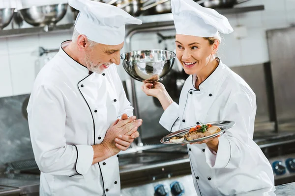 Female chef in uniform presenting meat dish on serving tray to male cook in restaurant kitchen — Stock Photo