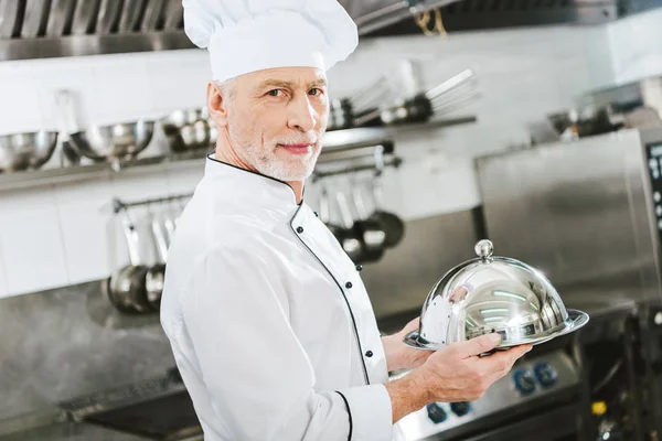 Guapo chef masculino en uniforme mirando a la cámara y sosteniendo bandeja de servir con cúpula en la cocina del restaurante - foto de stock