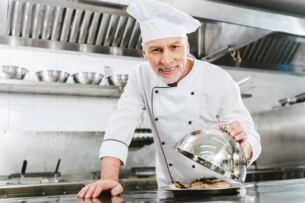 Bonito sorridente masculino chef no uniforme segurando cúpula de servir bandeja com prato de carne na cozinha do restaurante — Fotografia de Stock