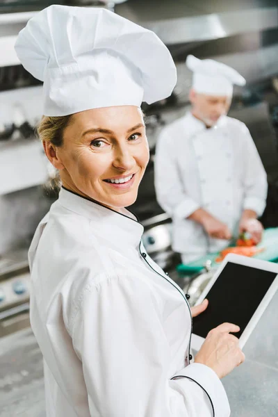 Selektiver Fokus der lächelnden Köchin, die in die Kamera schaut und in der Restaurantküche ein digitales Tablet mit leerem Bildschirm verwendet — Stockfoto