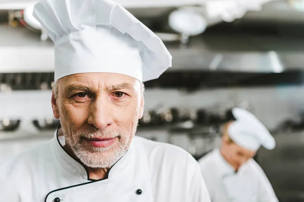 Beau chef masculin en uniforme et casquette regardant caméra dans la cuisine du restaurant avec espace de copie — Photo de stock