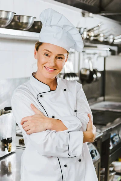 Привлекательная женщина-повар в форме с скрещенными руками, смотрящая в камеру на кухне ресторана — стоковое фото