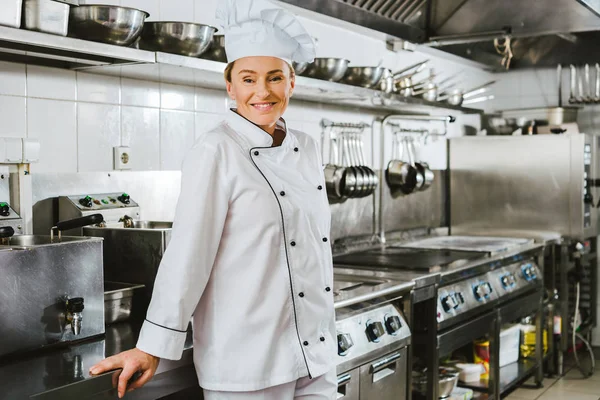 Attraktive lächelnde Köchin in Uniform und Hut, die in der Restaurantküche mit Kopierraum in die Kamera blickt — Stockfoto