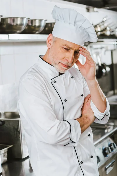 Tired male chef in uniform touching head and having headache in restaurant kitchen — Stock Photo
