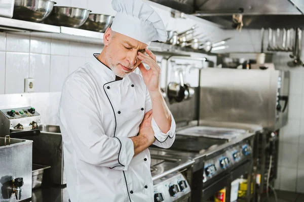Chef masculino cansado en uniforme tocando la cabeza y tener dolor de cabeza en la cocina del restaurante - foto de stock