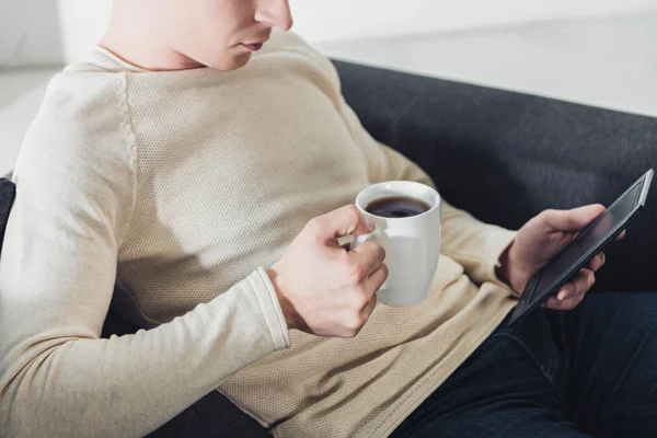 Vista recortada del hombre sosteniendo la taza con café y libro electrónico de lectura - foto de stock