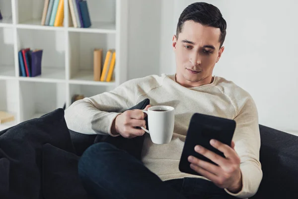 Uomo concentrato borchie con e-book mentre seduto sul divano a casa — Foto stock