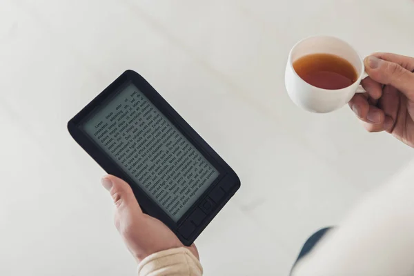 Abgeschnittene Ansicht von eBook und Tasse Tee in den Händen des Mannes zu Hause — Stockfoto